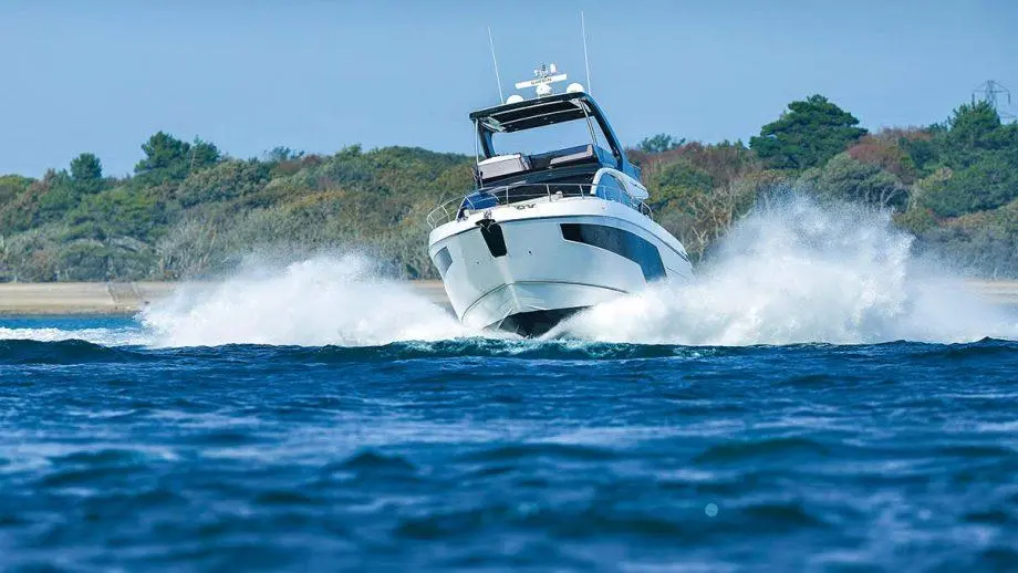 A luxury motorboat speeding through bright blue water, creating white waves, with greenery in the background.